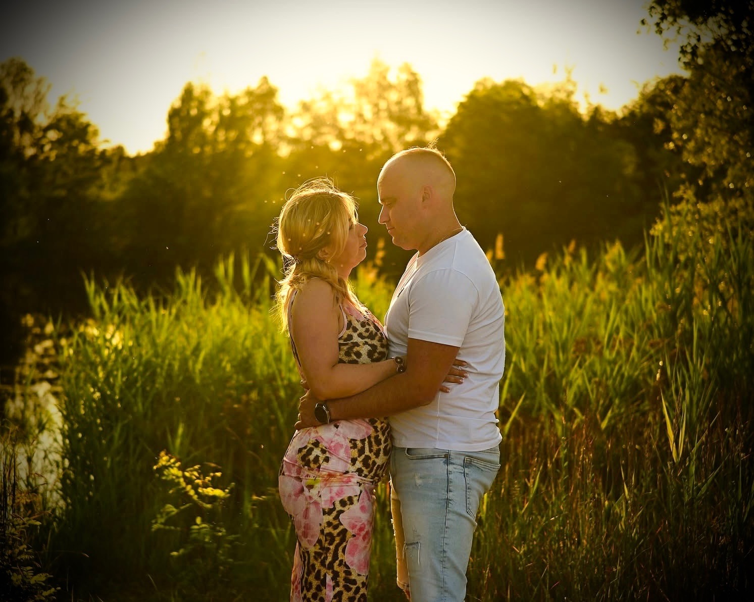 Zakochana para, kobieta w kwiecistej sukience ciążowej, mężczyzna w białej koszulce, obejmują się na tle zieleni i zachodzącego słońca. Romantyczna sesja w plenerze.