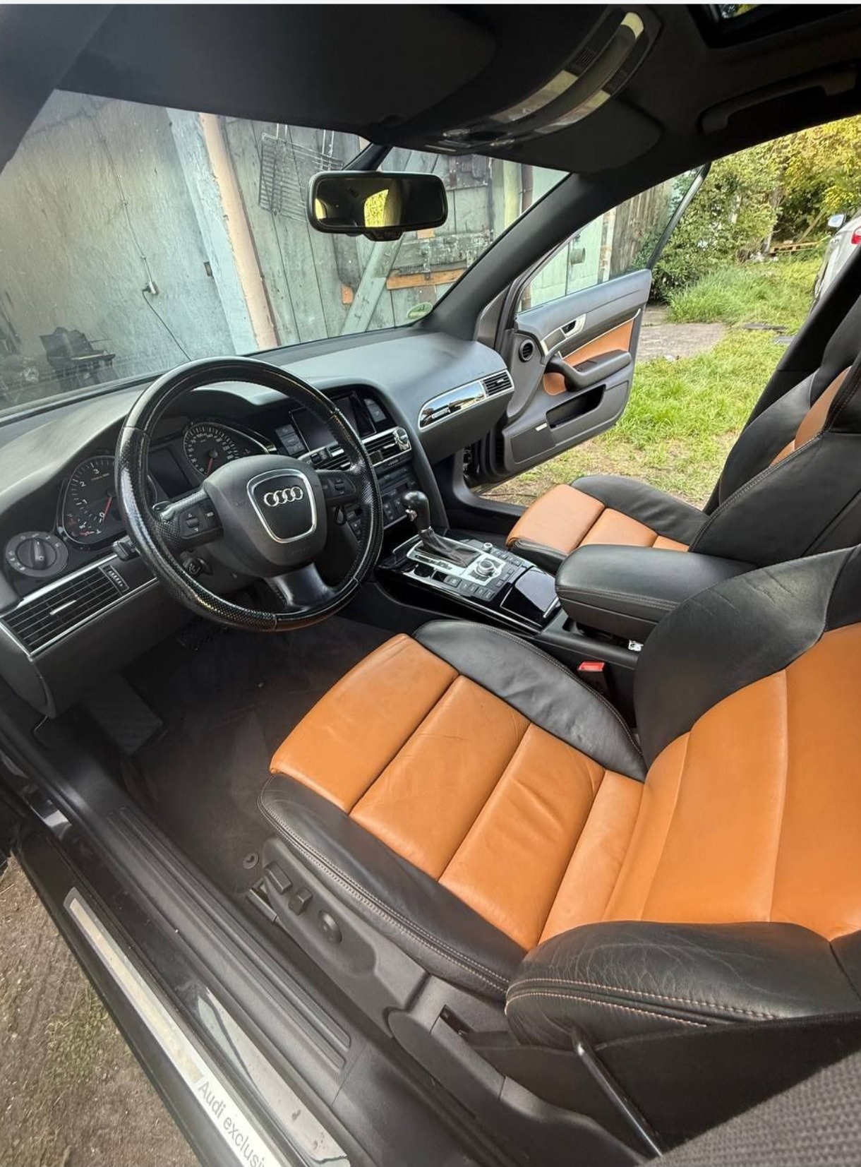 Wnętrze czarnego Audi A6 z brązowo-czarną skórzaną tapicerką, widok z zewnątrz przez otwarte drzwi, ukazujący deskę rozdzielczą i fotele.