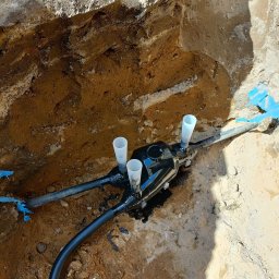 Usługi elektryczne - Wykop z ziemi z czarną puszką i rurami, prawdopodobnie przyłącze elektryczne, z trzema białymi kielichami na górze, w ziemi.