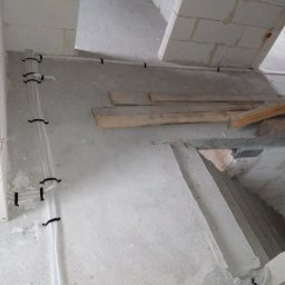 Usługi elektryczne - Białe, karbowane rury elektryczne przymocowane czarnymi uchwytami do surowej, betonowej ściany w trakcie budowy. Widoczne bloki betonowe i drewniane elementy konstrukcyjne.