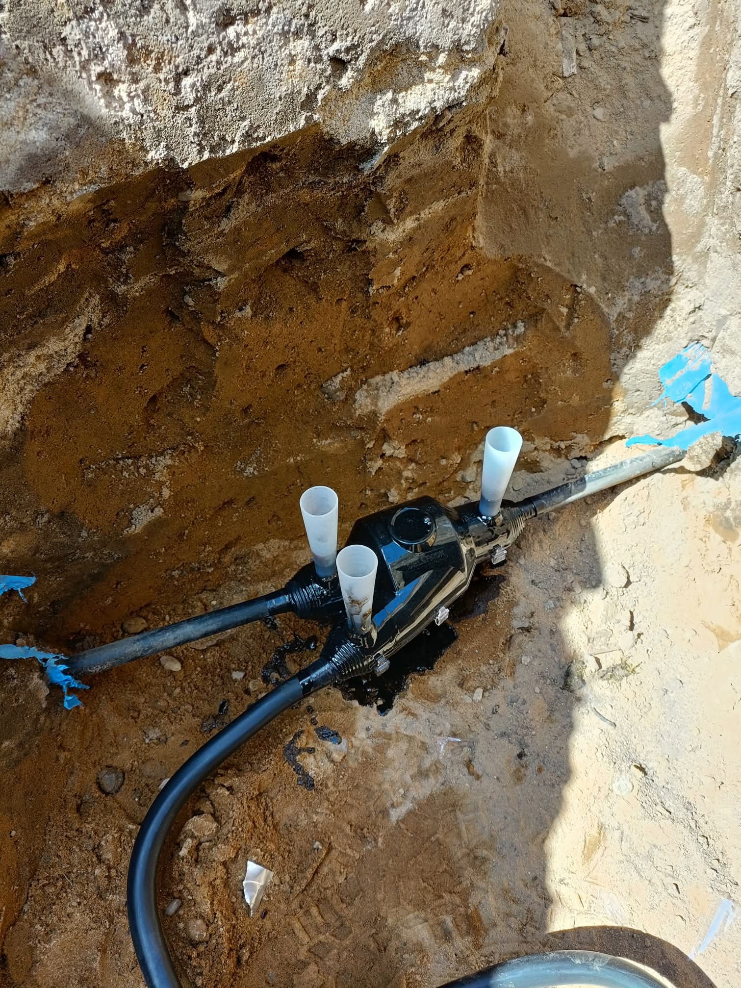 Wykop z ziemi z czarną puszką i rurami, prawdopodobnie przyłącze elektryczne, z trzema białymi kielichami na górze, w ziemi.