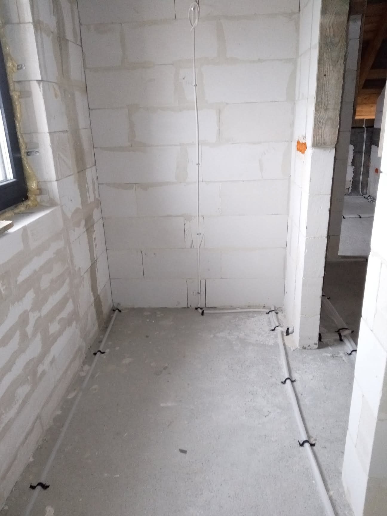 Wnętrze w trakcie prac elektrycznych: przewody ułożone wzdłuż ścian, pionowe kable przytwierdzone do ściany z betonu komórkowego. Widoczne okno i surowy stan wykończenia.