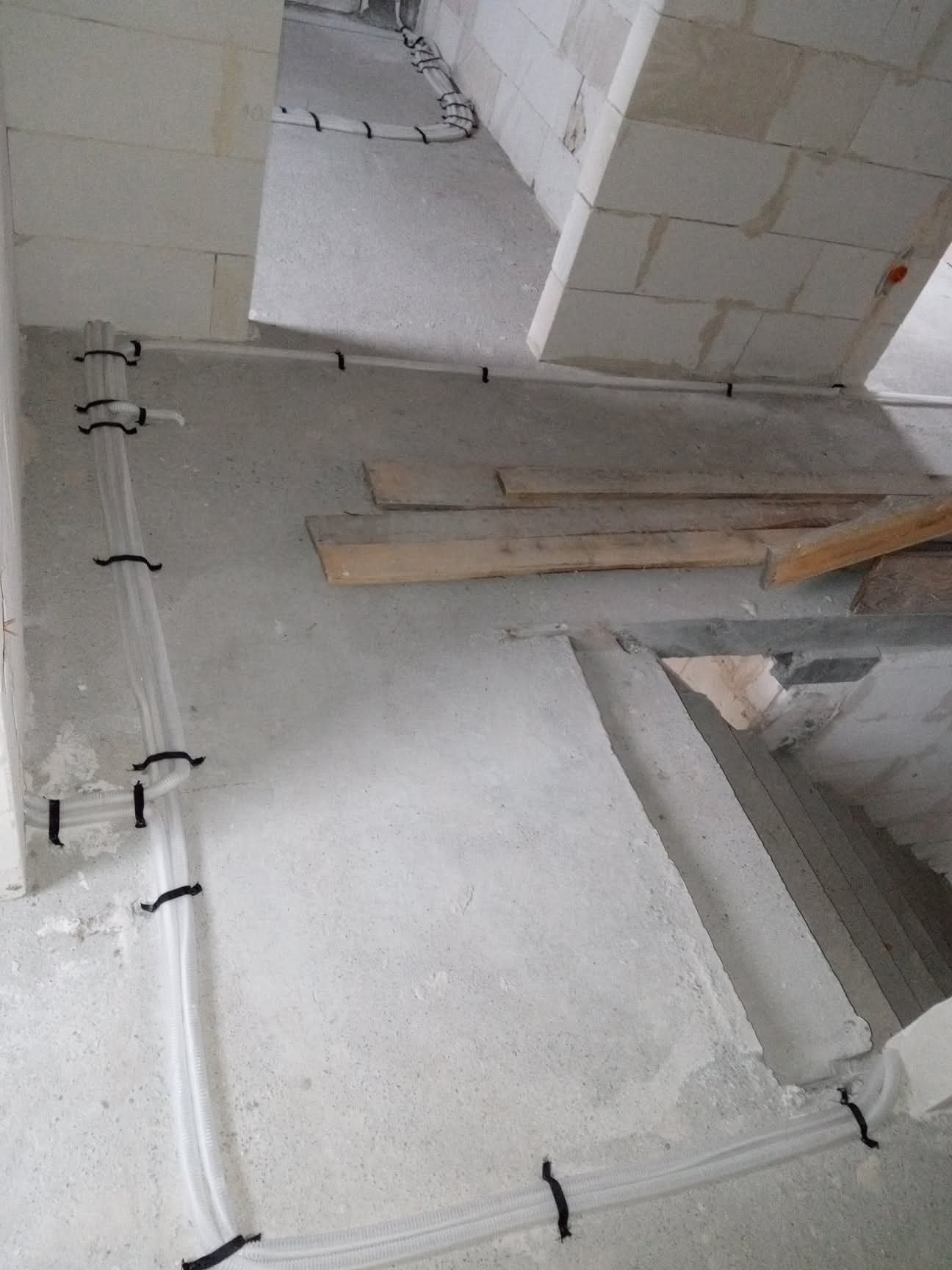 Białe, karbowane rury elektryczne przymocowane czarnymi uchwytami do surowej, betonowej ściany w trakcie budowy. Widoczne bloki betonowe i drewniane elementy konstrukcyjne.