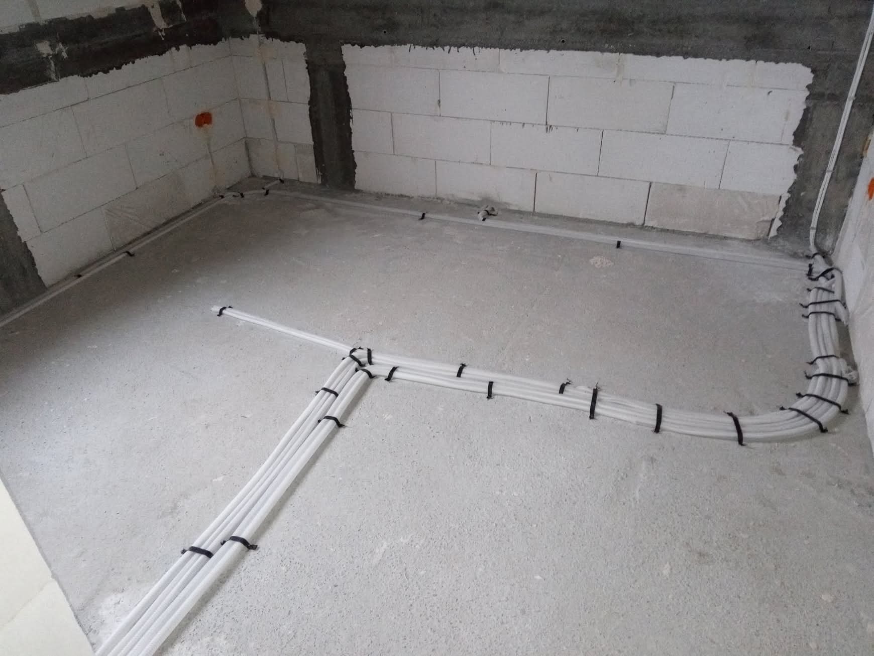 Białe rury instalacji elektrycznej ułożone na surowym betonie wzdłuż ścian z białego bloczka, zabezpieczone czarnymi opaskami zaciskowymi. Widok z góry, surowy stan.