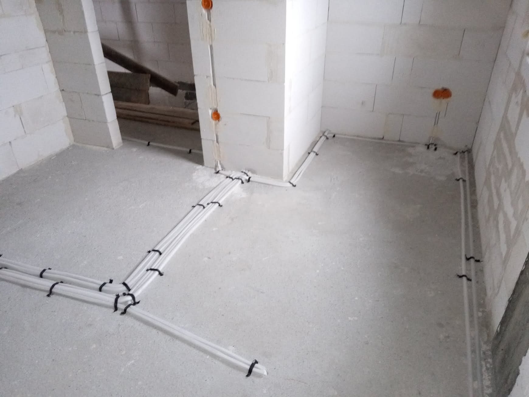 Białe rury instalacji elektrycznej zamocowane do surowej posadzki w pomieszczeniu z białymi ścianami z betonu komórkowego. Widoczne pomarańczowe puszki elektryczne.
