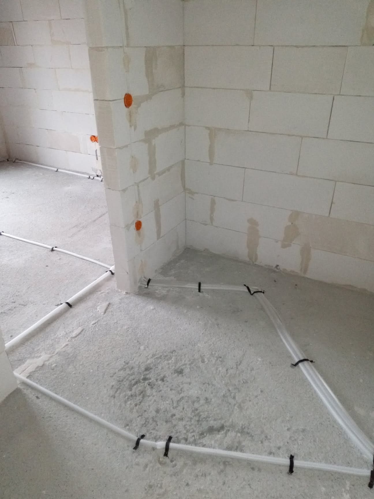 Surowy stan: białe rury instalacji grzewczej podłogowej, przymocowane czarnymi uchwytami do szarego betonu w pomieszczeniu z surowymi ścianami z białych bloczków.