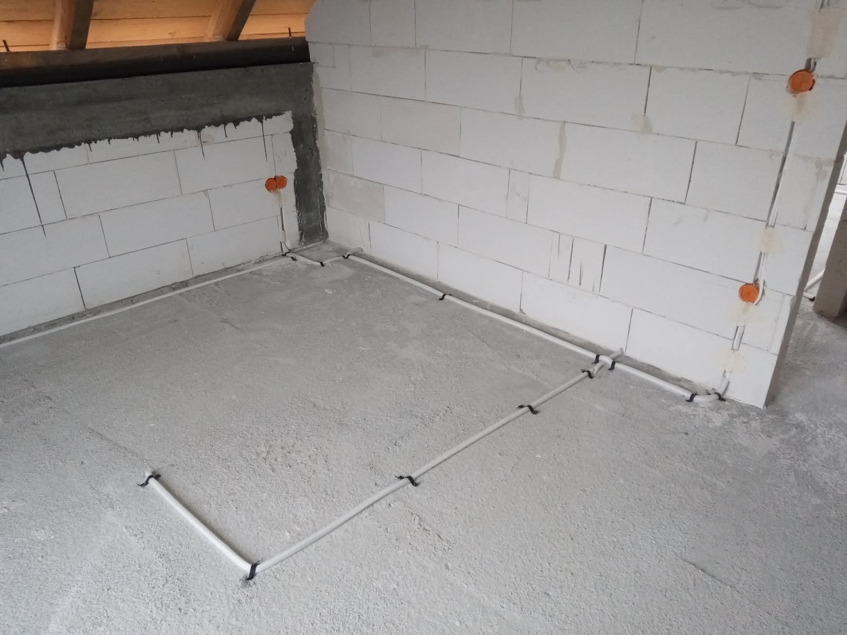 Surowy stan: białe ściany z betonu komórkowego, podłoga wylewka, rury elektryczne poprowadzone po podłodze i w ścianach, pomarańczowe puszki elektryczne.