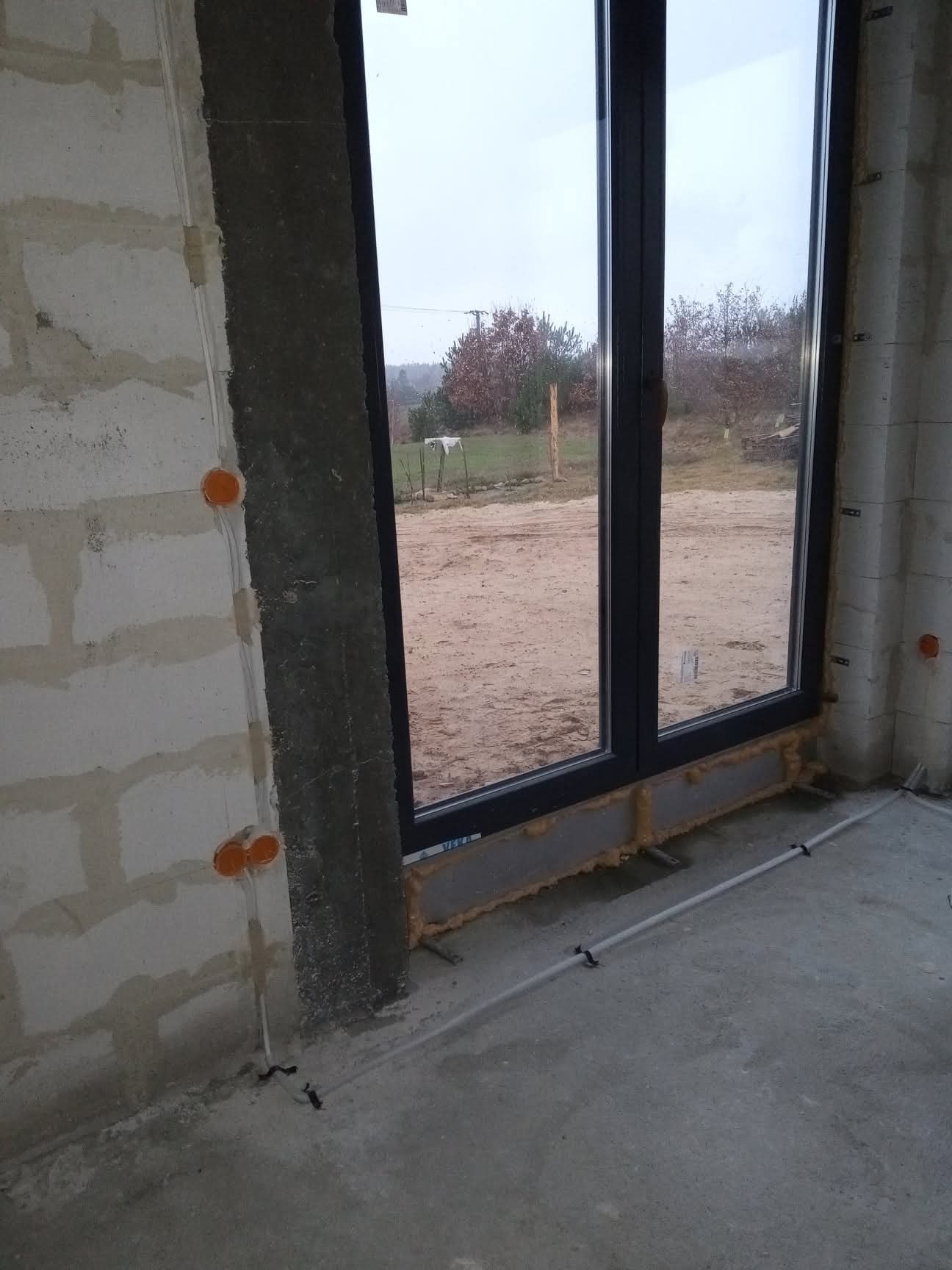 Surowy stan: okno PCV w ścianie z betonu komórkowego, widoczne puszki elektryczne i przewody. W tle widok na zewnątrz przez okno.