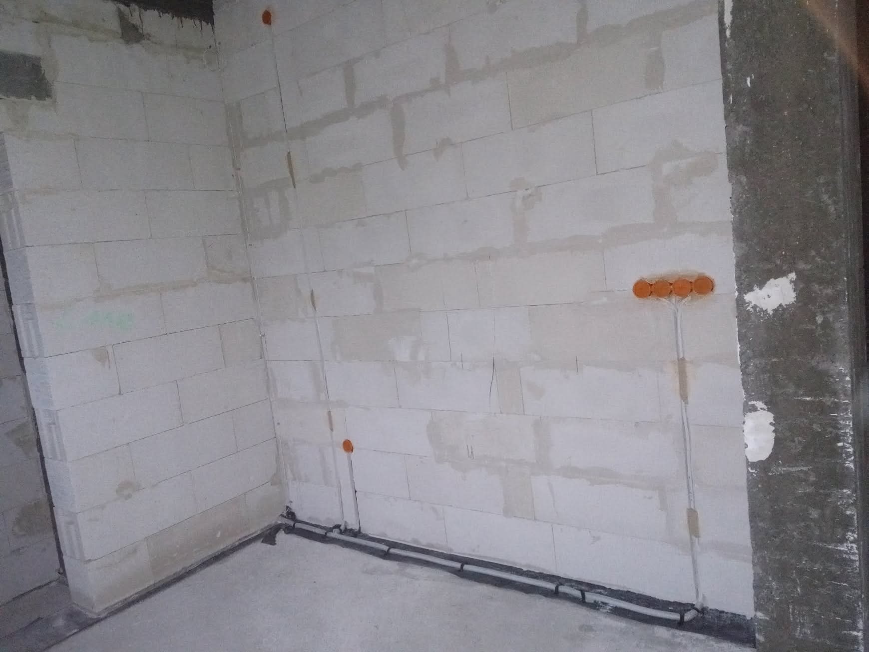 Ściana z betonu komórkowego z widoczną instalacją elektryczną: rurki ochronne i pomarańczowe puszki pod gniazdka. Surowy stan, przygotowanie do dalszych prac.