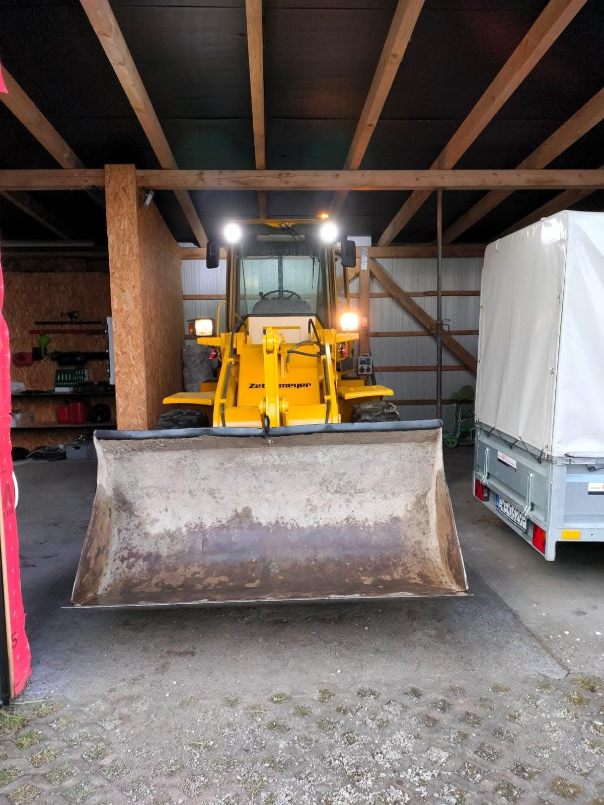 Żółta ładowarka Zet Meyer z podniesioną łyżką, stojąca w zadaszonym miejscu z drewnianą konstrukcją i przyczepą w tle. Widok na kabinę operatora.