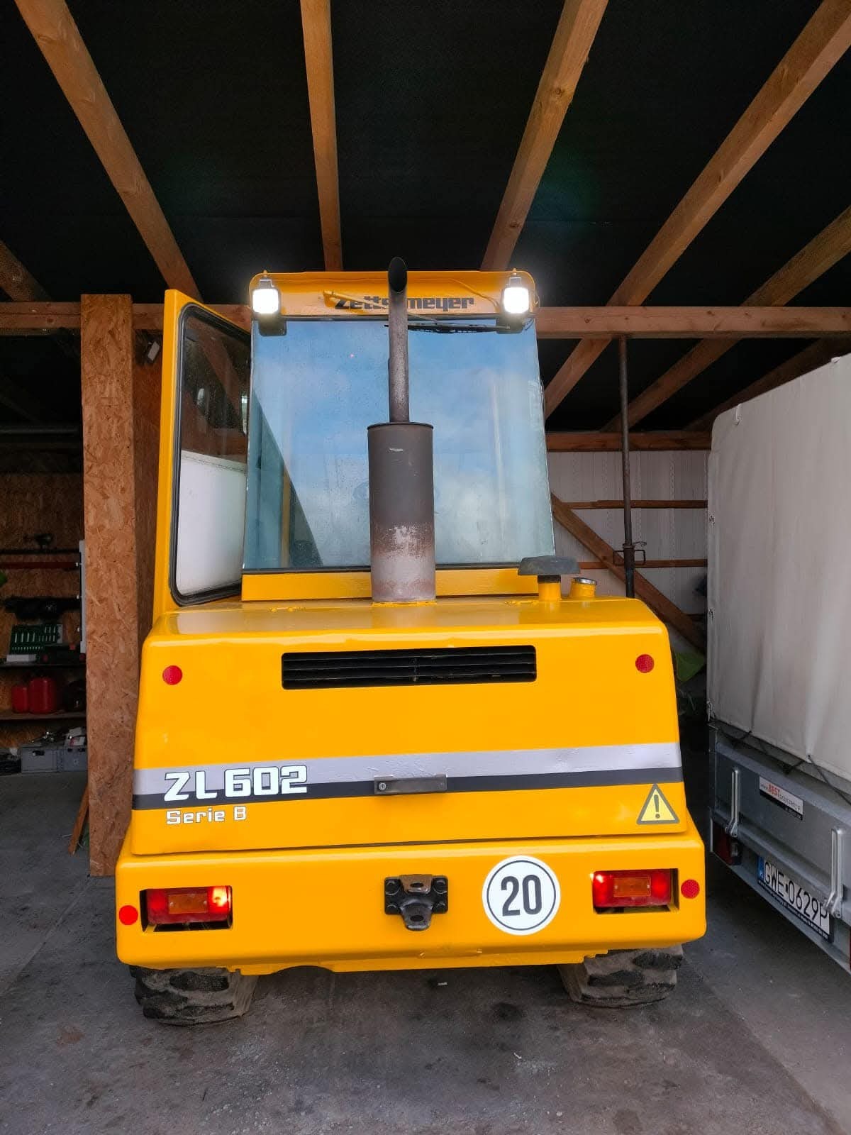 Żółty ciągnik Zettelmeyer ZL602 Serie B z kabiną i oświetleniem, widoczny z tyłu, stojący w zadaszonym pomieszczeniu z drewnianą konstrukcją sufitu i przyczepką obok.