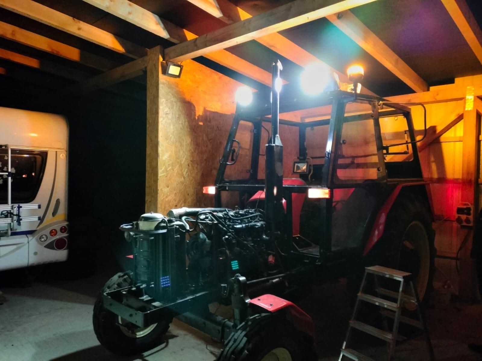 Czerwony traktor z włączonymi światłami, zaparkowany w zadaszonym garażu obok białej przyczepy kempingowej, oświetlony ciepłym światłem żarówek.
