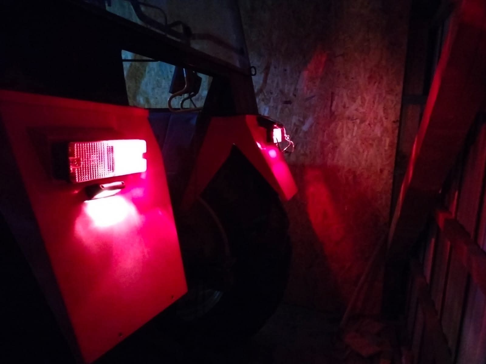 Czerwone światło lampy zamontowanej na pojeździe, oświetlające drewnianą ścianę. Widoczne elementy konstrukcyjne pojazdu i opona w tle.