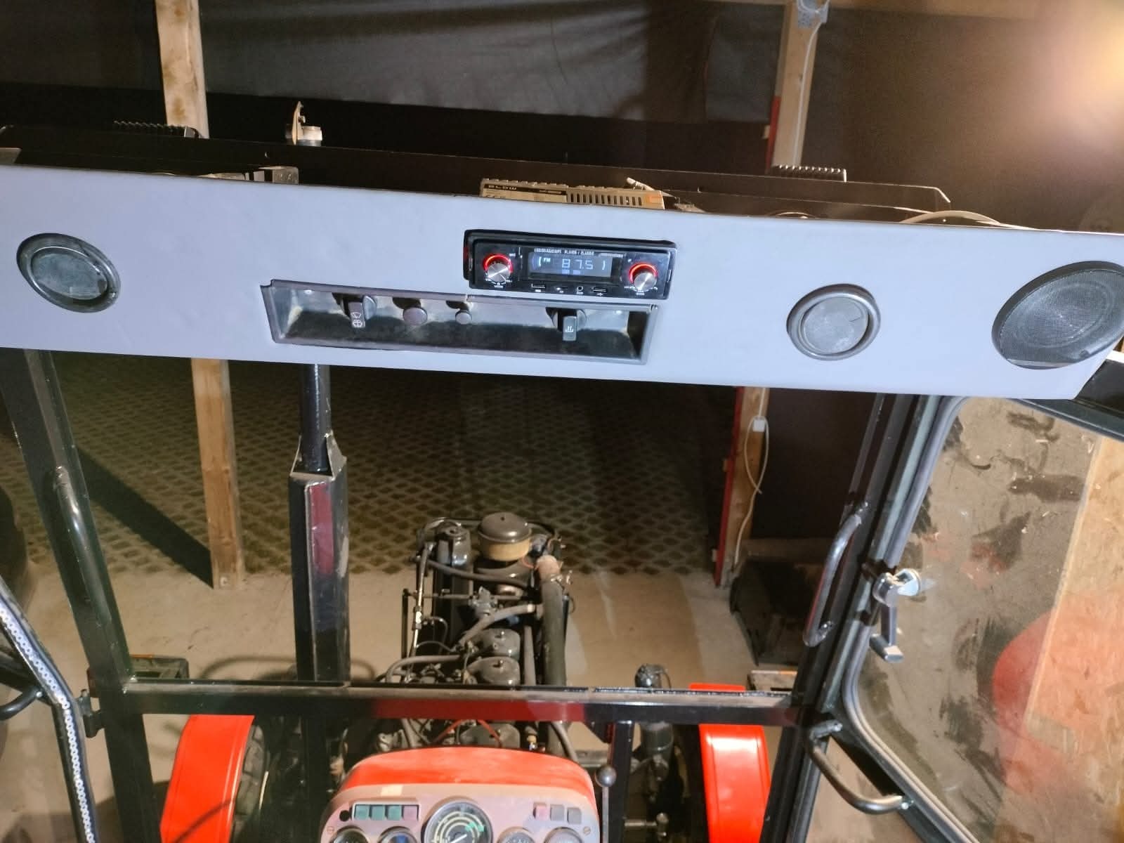 Wnętrze kabiny traktora z zamontowanym radiem samochodowym i głośnikami w szarej obudowie podsufitowej. Widok z perspektywy kierowcy na deskę rozdzielczą i silnik.