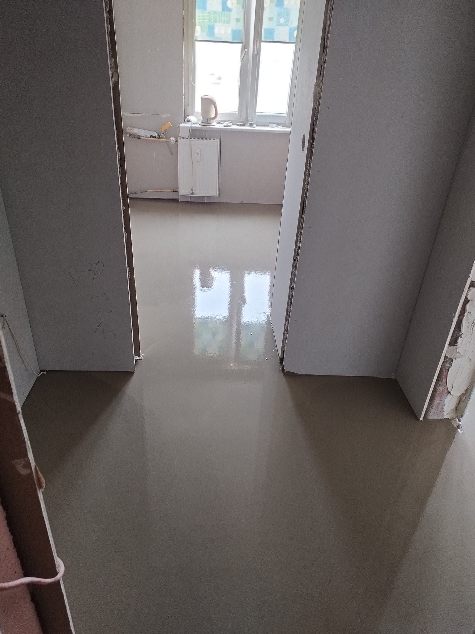 Wylana posadzka samopoziomująca w odcieniach szarości, widoczna przez futryny drzwi. W tle okno z parapetem i grzejnikiem.