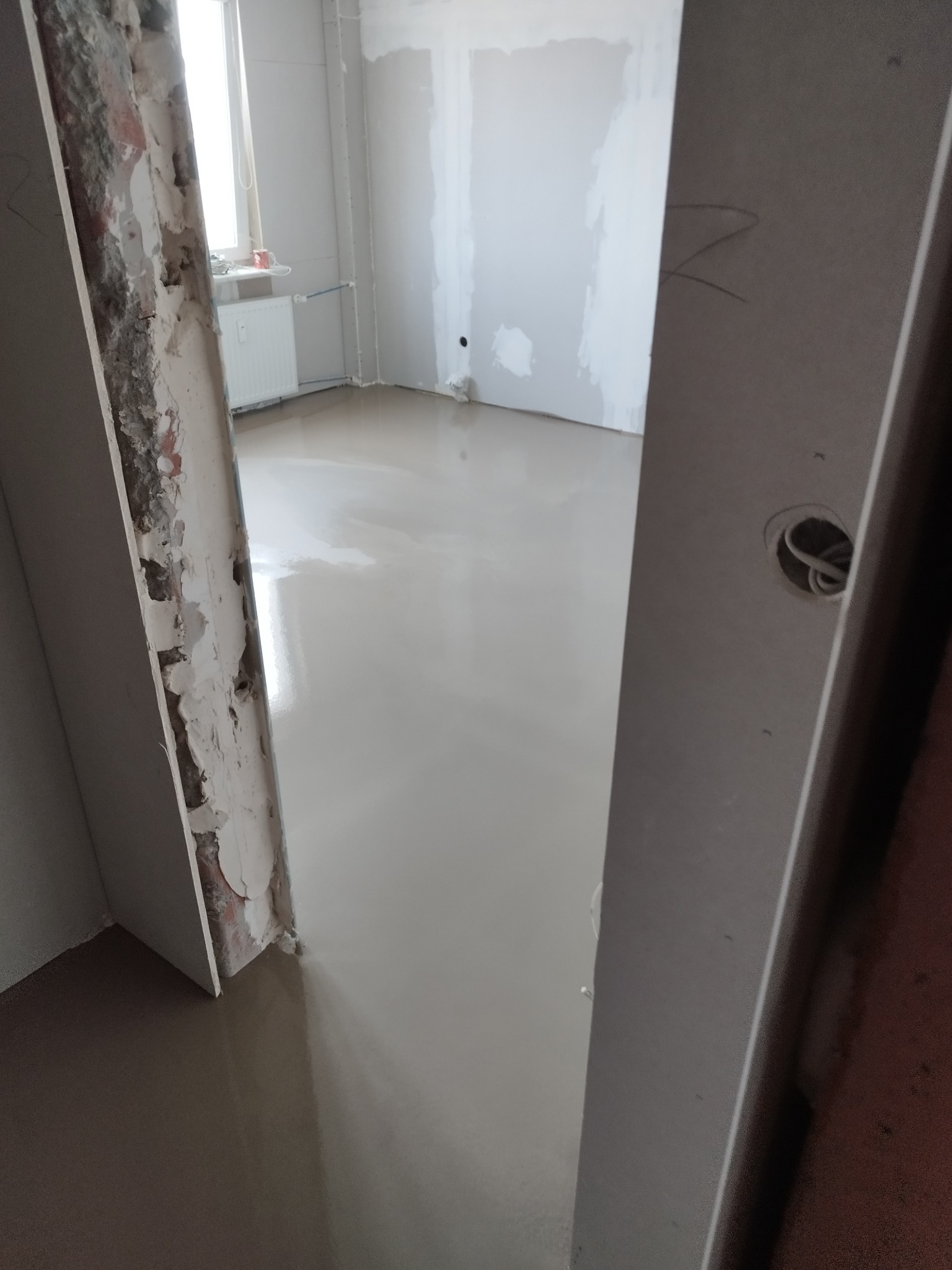 Wylany, gładki podkład podłogowy w jasnym pomieszczeniu w trakcie remontu. Widoczne ślady po usuniętych płytkach przy framudze drzwi i przygotowane ściany.