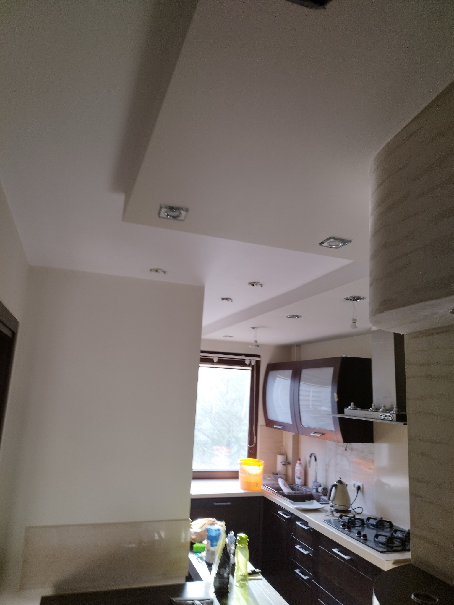 Jasna kuchnia po malowaniu: świeżo pomalowany sufit z oświetleniem punktowym, kremowe ściany, ciemne meble kuchenne, okno z widokiem na zewnątrz.