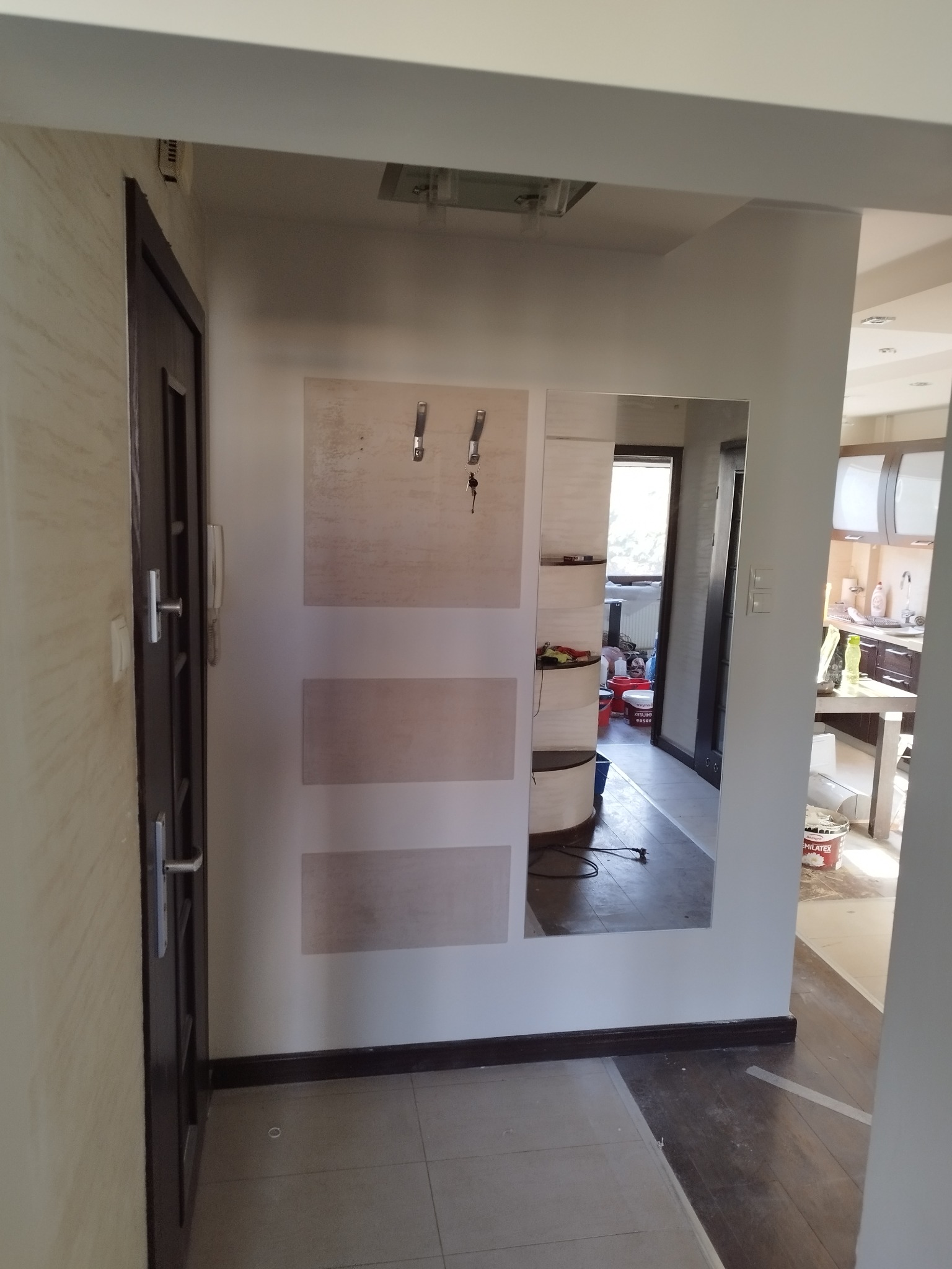 Przedpokój z lustrem w zabudowie i widocznym odbiciem wnętrza mieszkania. Na podłodze płytki, ściany pomalowane na jasny kolor. Widoczny fragment drzwi.