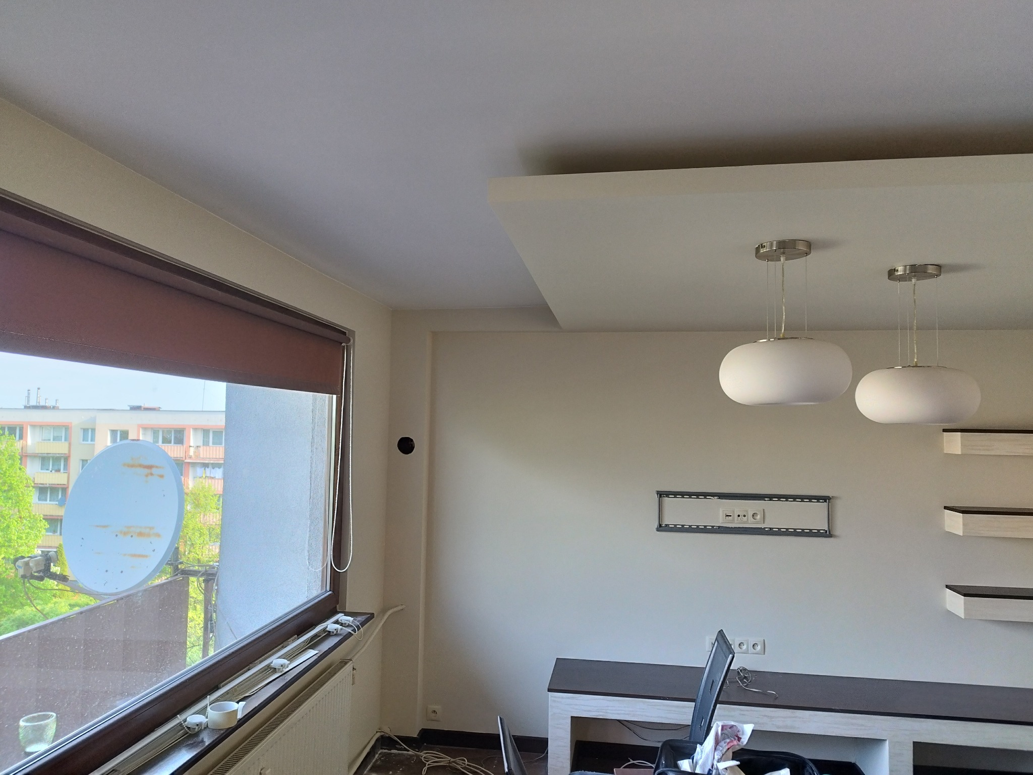 Jasne wnętrze po remoncie: białe ściany, designerskie lampy, zamontowany uchwyt TV, półki. Widok z okna na satelitę. Minimalistyczny design i czysta przestrzeń.