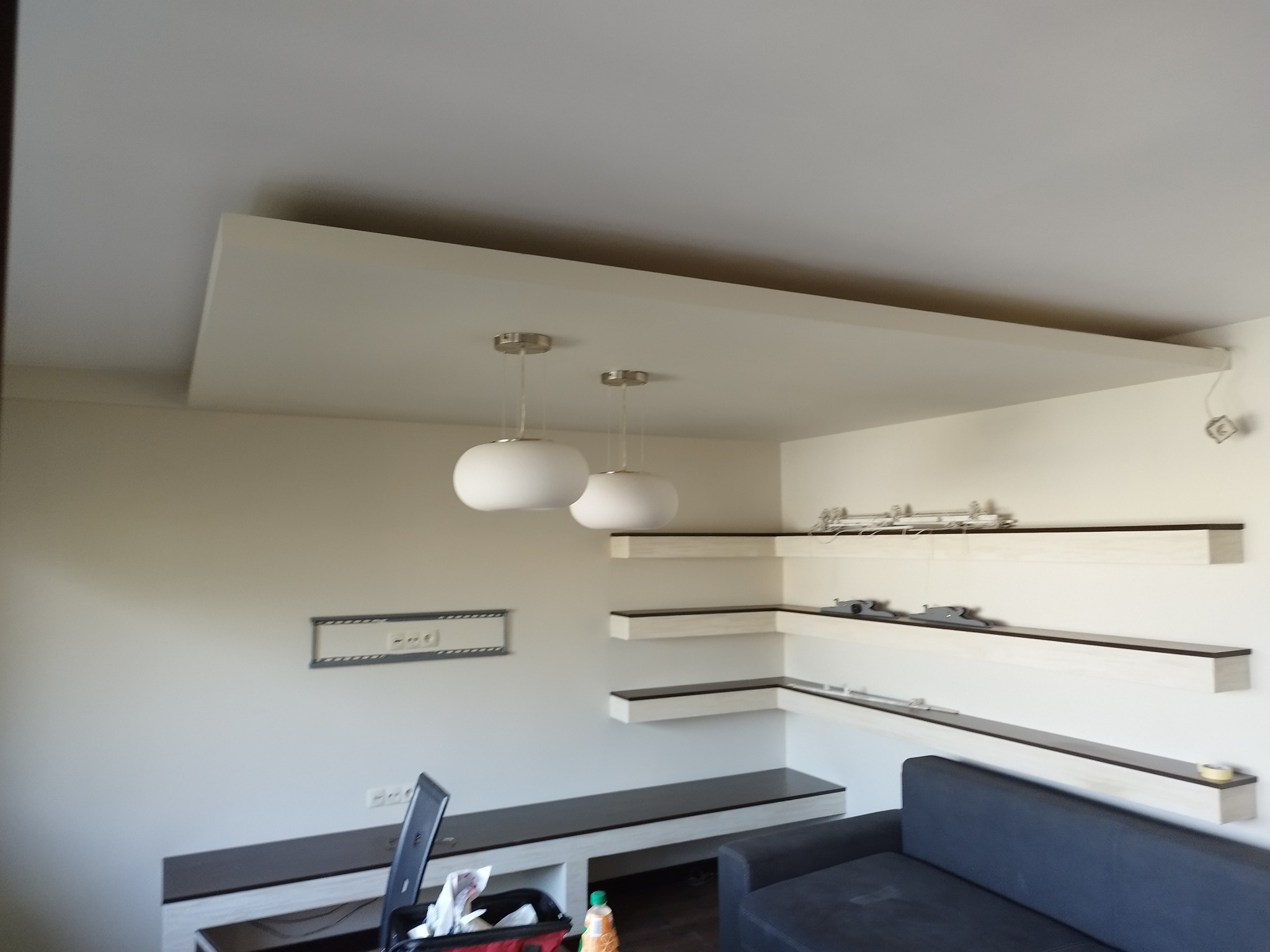 Nowoczesne wnętrze: podwieszany sufit z oświetleniem, minimalistyczne półki narożne i ciemna sofa. Widoczne gniazdka i elementy instalacji.