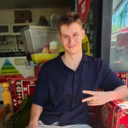 Młody mężczyzna siedzi na wzorzystym fotelu na tle baru z sokami, uśmiechając się i pokazując gest 'V'.