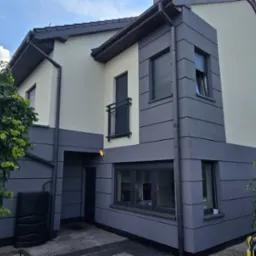 Nowoczesny dom z dwukolorową elewacją w odcieniach szarości i beżu, z widocznymi detalami architektonicznymi i zielenią w tle, uchwycony w słoneczny dzień.