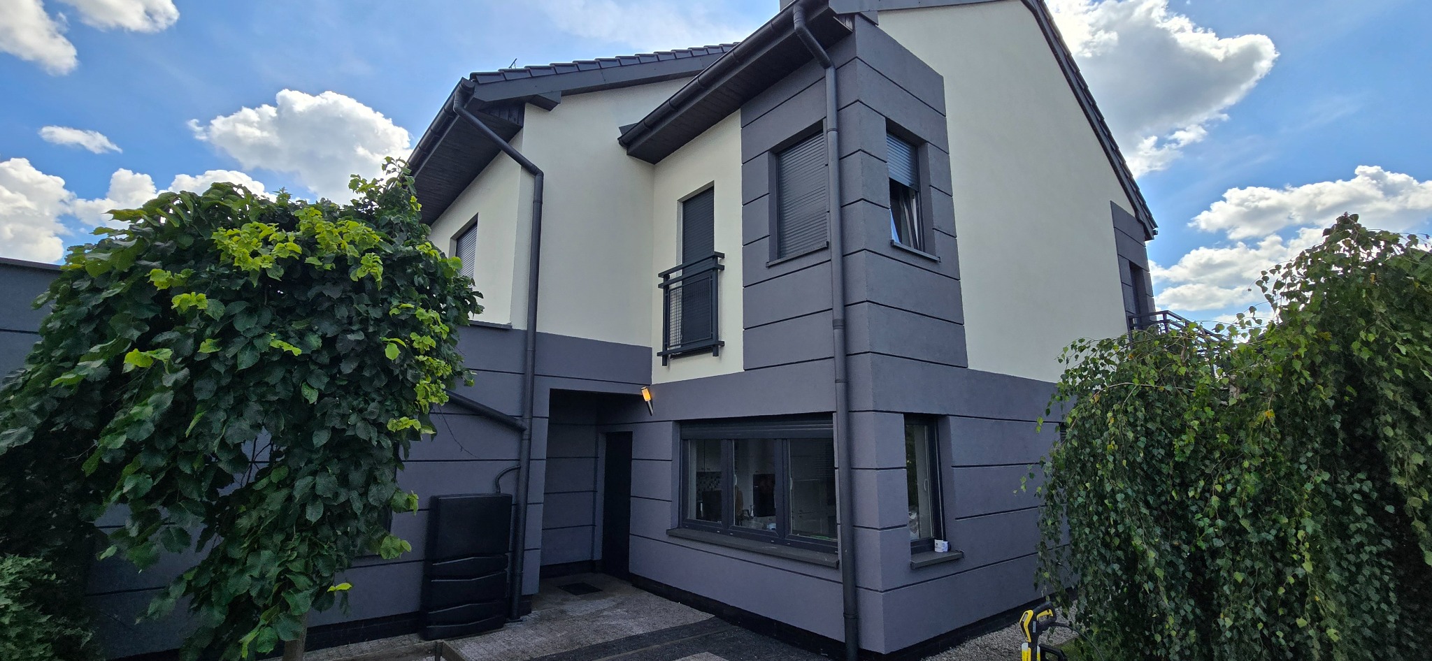 Nowoczesny dom z dwukolorową elewacją w odcieniach szarości i beżu, z widocznymi detalami architektonicznymi i zielenią w tle, uchwycony w słoneczny dzień.