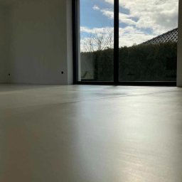 ACH DECOR - Jasna, gładka posadzka z mikrocementu w nowoczesnym wnętrzu z dużym oknem na ogród. Minimalistyczny design, naturalne światło.