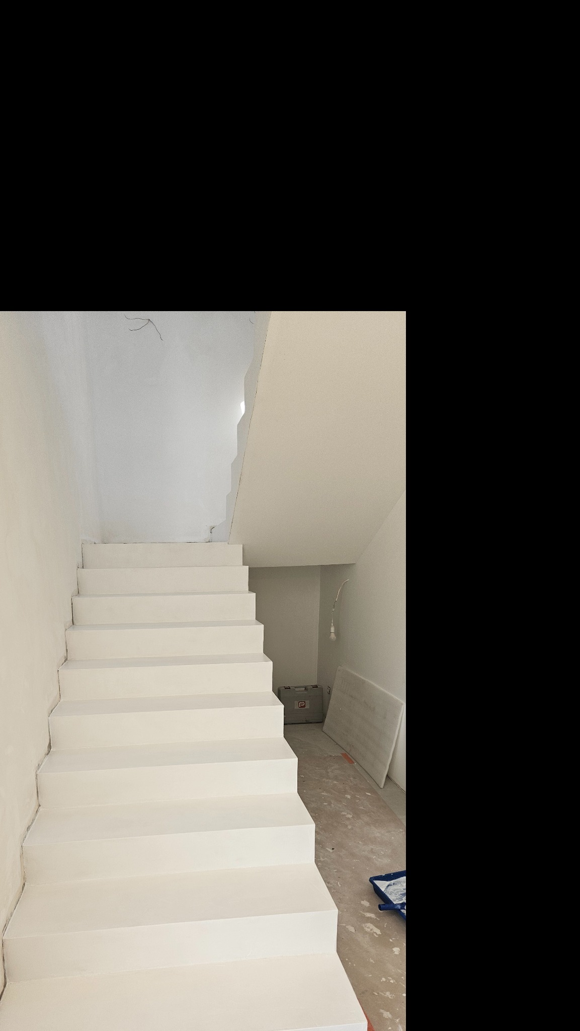 Białe schody w trakcie malowania w jasnym wnętrzu. Widoczne narzędzia malarskie i niedomalowane fragmenty ścian.