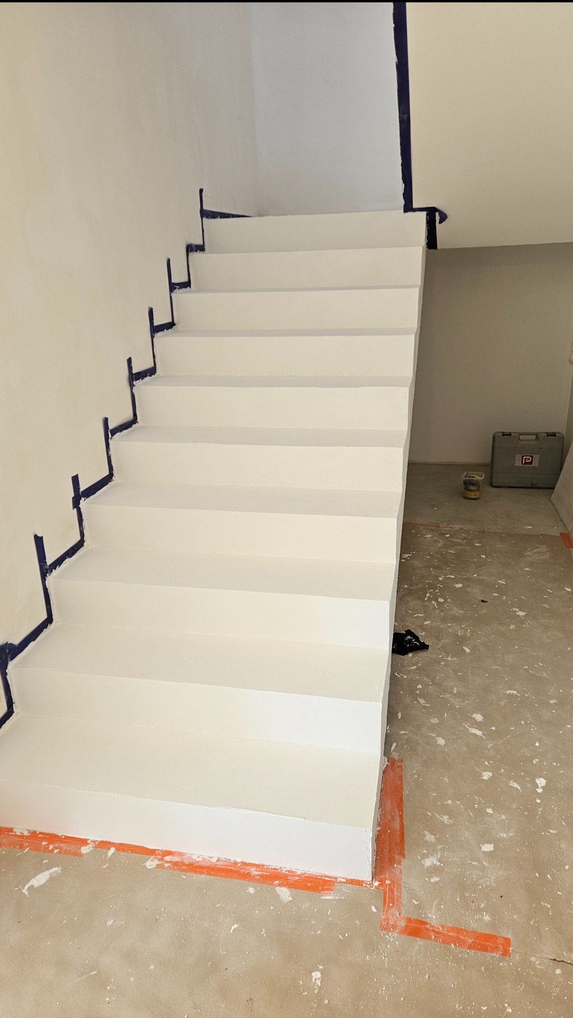 Białe schody z zabezpieczeniami malarskimi, częściowo pomalowane, widoczne narzędzia malarskie w tle, remont w trakcie.
