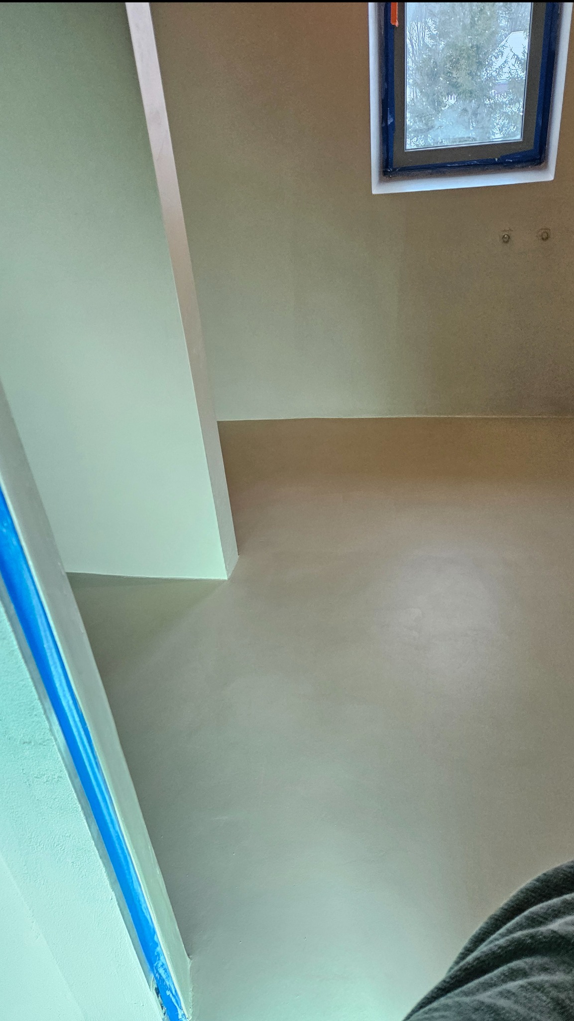 Ściana i sufit pokryte gładzią mikrocementową, z zabezpieczonym oknem taśmą malarską. Widoczne detale wykończenia i faktura materiału.