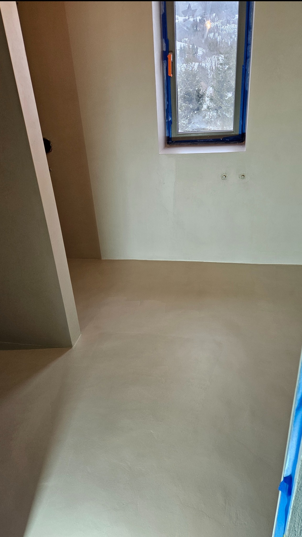 Jasne wnętrze po wykonaniu wylewki, widoczne okno z widokiem na ośnieżone drzewa. Ściany w jasnym odcieniu. W rogu widoczny fragment pionowej konstrukcji.