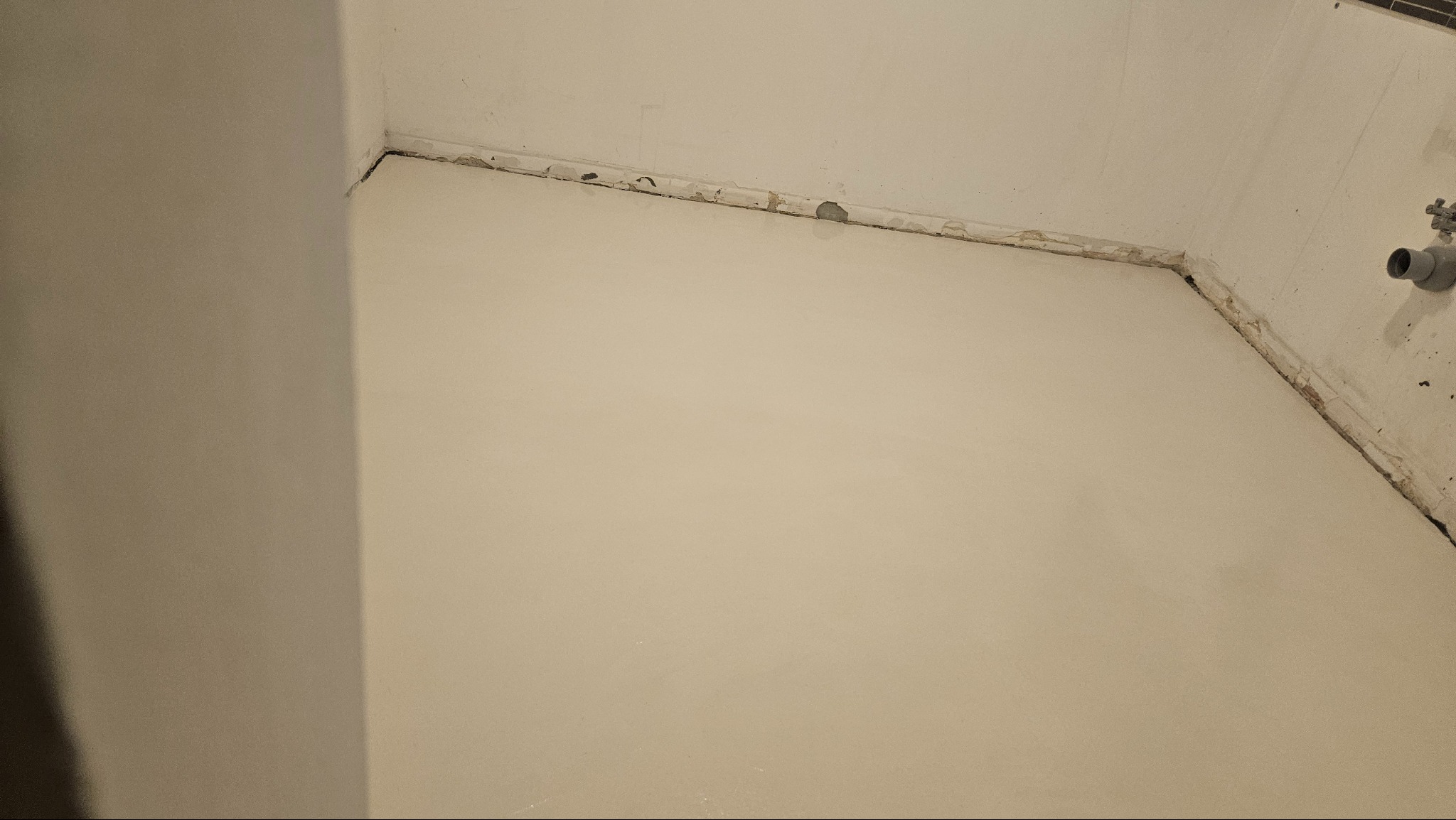 Gładka, beżowa ściana po renowacji w Łętowni, widok z dołu. Nierówności przy suficie, rura kanalizacyjna w rogu. Minimalistyczne wykończenie wnętrza.