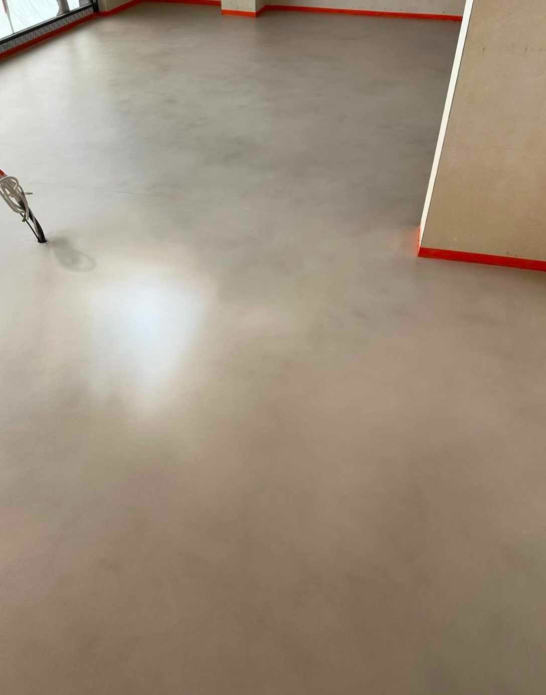 Jasna, jednolita posadzka z mikrocementu w nowym pomieszczeniu z widocznymi listwami przypodłogowymi i narzędziem do aplikacji. Gładka powierzchnia.