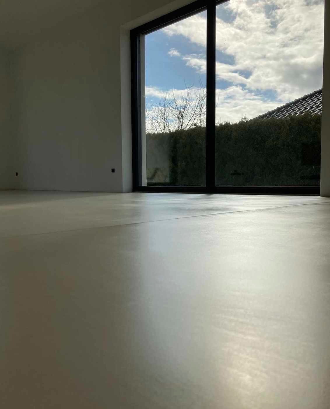 Jasna, gładka posadzka z mikrocementu w nowoczesnym wnętrzu z dużym oknem na ogród. Minimalistyczny design, naturalne światło.