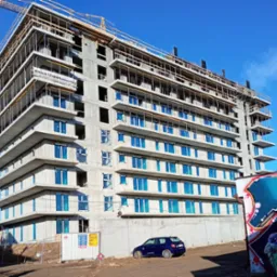 Wysoki budynek apartamentowy w trakcie budowy, z widocznym żurawiem i zabezpieczonymi oknami folią. Na dole zaparkowany niebieski samochód osobowy.