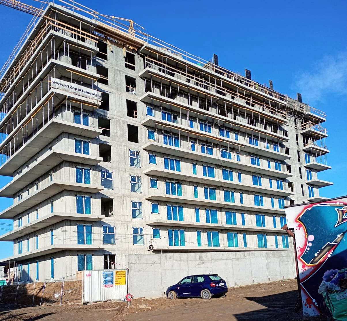 Wysoki budynek apartamentowy w trakcie budowy, z widocznym żurawiem i zabezpieczonymi oknami folią. Na dole zaparkowany niebieski samochód osobowy.