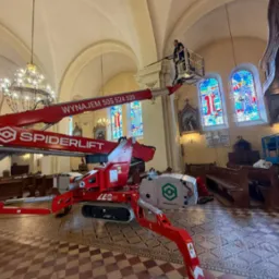Kościół w trakcie prac renowacyjnych: pracownik na podnośniku Spiderlift przy witrażach, widoczne ławki i żyrandol. Renowacja wnętrza sakralnego.
