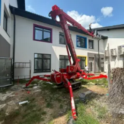 Prace elewacyjne z użyciem podnośnika Spiderlift. Biała fasada budynku z kolorowymi obramowaniami okien. Widoczny pracownik w koszu podnośnika.