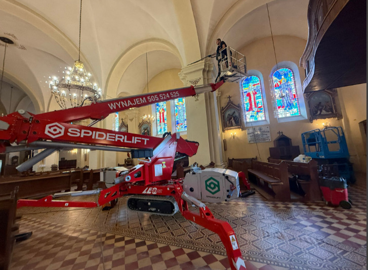 Kościół w trakcie prac renowacyjnych: pracownik na podnośniku Spiderlift przy witrażach, widoczne ławki i żyrandol. Renowacja wnętrza sakralnego.