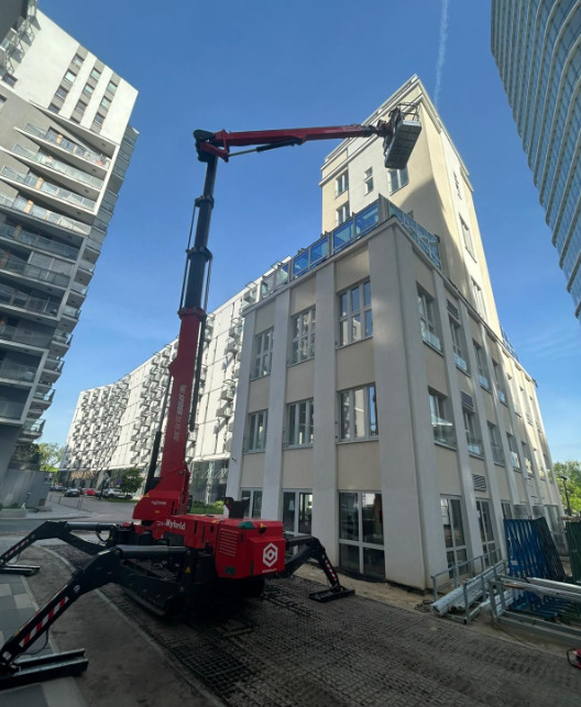 Podnośnik koszowy pająk na chodniku, na tle budynku z jasną elewacją. Maszyna gotowa do prac na wysokościach, pomiędzy blokami mieszkalnymi.