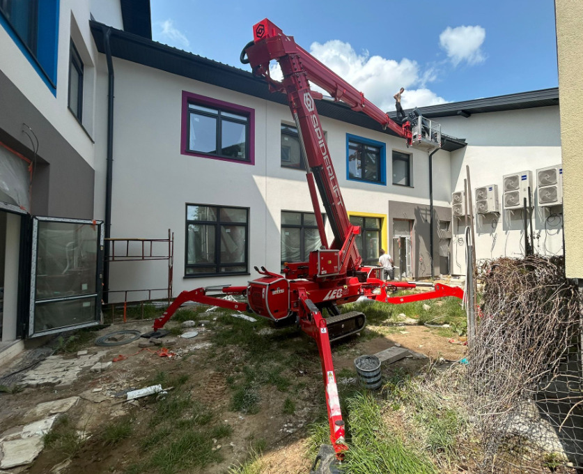 Prace elewacyjne z użyciem podnośnika Spiderlift. Biała fasada budynku z kolorowymi obramowaniami okien. Widoczny pracownik w koszu podnośnika.