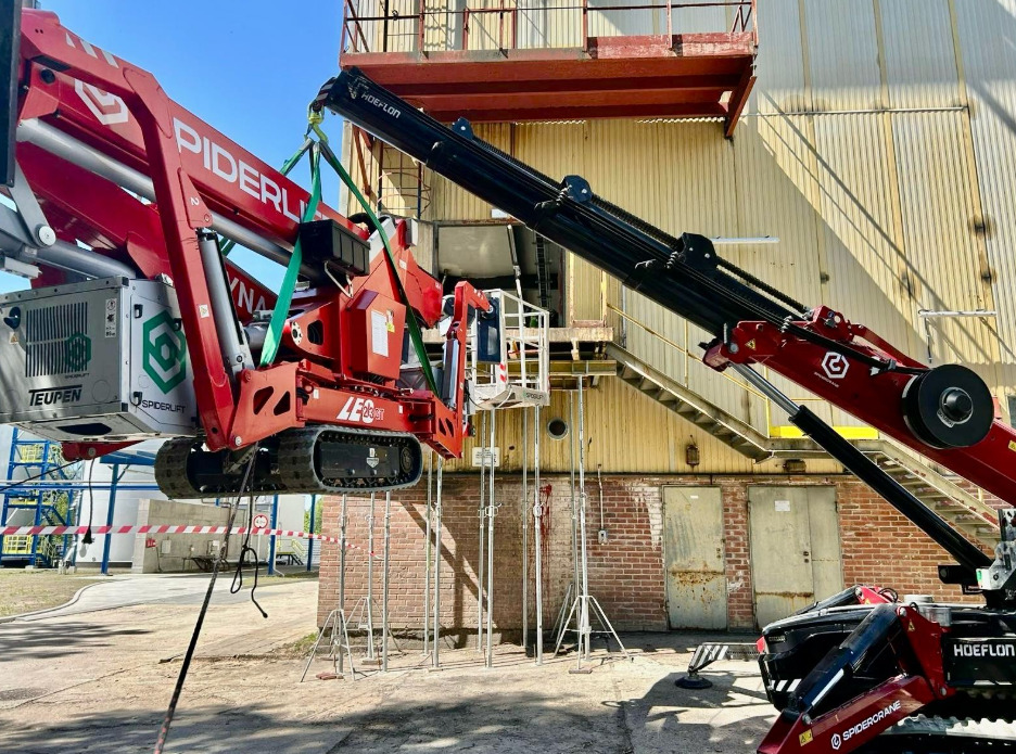 Czerwony podnośnik koszowy Spiderlift na gąsienicach, wsparty na podporach, przy żółtej elewacji budynku z ceglanym dołem, przygotowany do prac na wysokości.