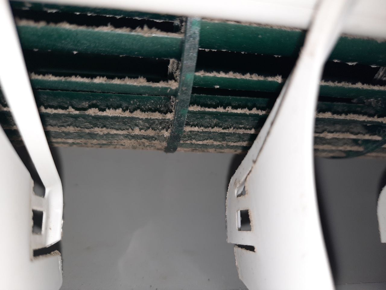 Zanieczyszczone, zielone lamele klimatyzatora z widocznym nagromadzonym kurzem, uchwycone z bliskiej perspektywy. Białe elementy obudowy na pierwszym planie.