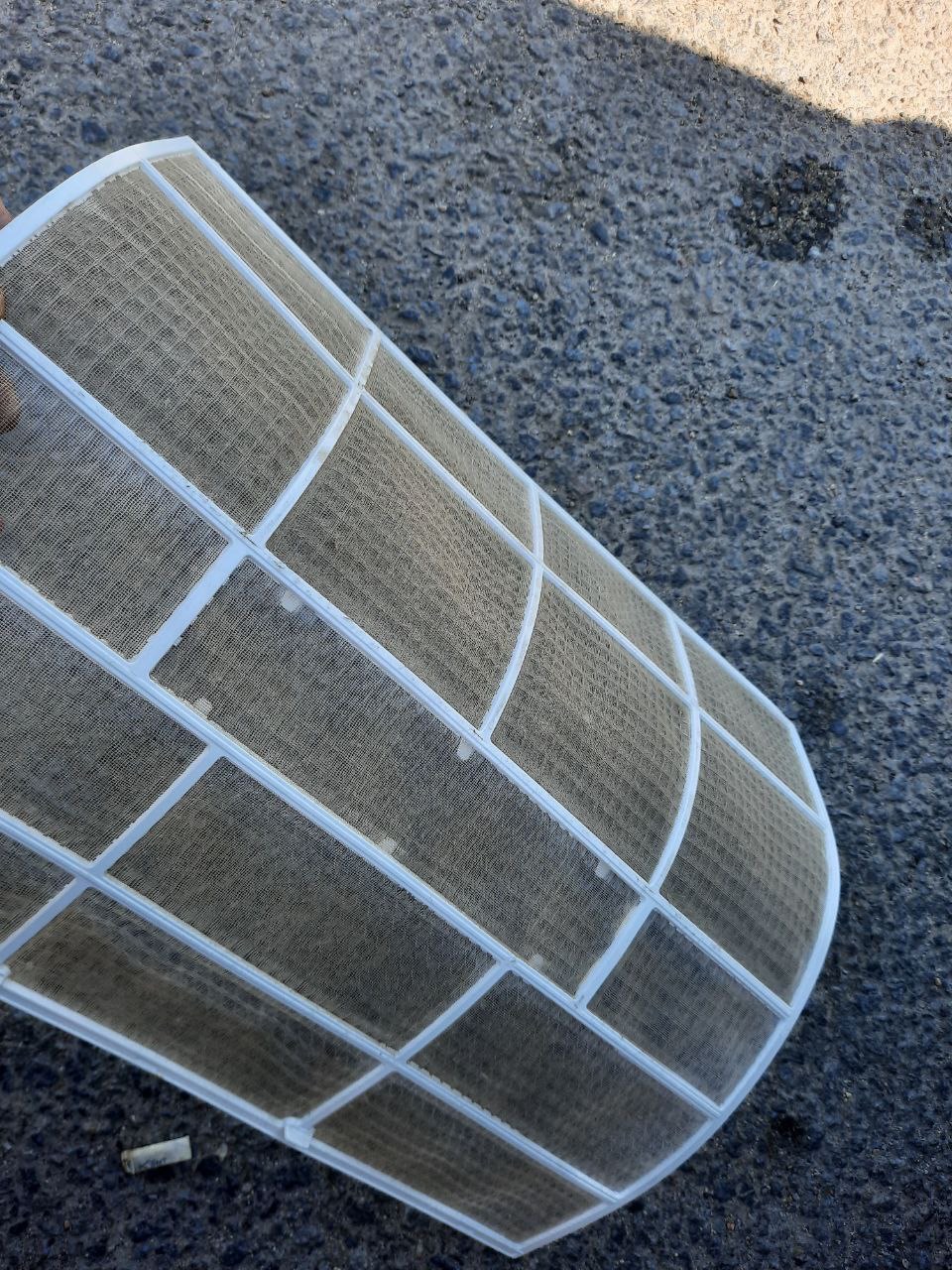 Brudny filtr klimatyzacji z widocznymi zanieczyszczeniami leżący na szorstkiej, ciemnej powierzchni. Filtr jest biały z siatką.