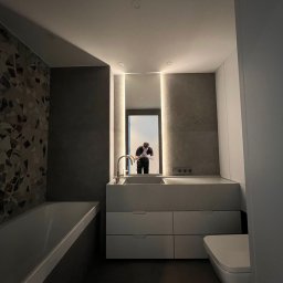 BluMeble - Nowoczesna łazienka z umywalką w zabudowie, minimalistyczną toaletą i mozaiką na ścianie. W lustrze odbija się osoba. Stonowana kolorystyka i oświetlenie LED.