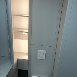 BluMeble - Szara szafa wnękowa z oświetleniem wewnętrznym, otwarte drzwi ukazują puste półki. Widoczny włącznik światła i fragment toalety.