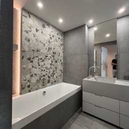 BluMeble - Nowoczesna łazienka z wanną obudowaną szarą mozaiką, umywalką z szafką w odcieniach szarości i dużym lustrem odbijającym fragment wnętrza.