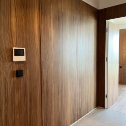 BluMeble - Ściana z forniru orzechowego z ukrytymi drzwiami i szafami, minimalistyczny design, biała listwa przypodłogowa i sufitowa, wideodomofon na ścianie.