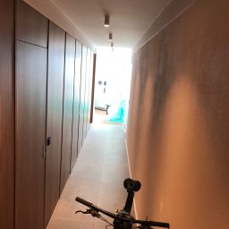 BluMeble - Długi korytarz z zabudową szaf w odcieniach brązu i beżowymi ścianami. W tle widoczne jasne pomieszczenie. Na pierwszym planie fragment roweru.